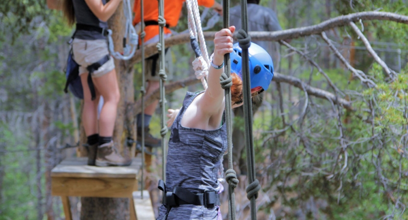A student wearing safety gear is secured by ropes as they navigate a ropes course. 